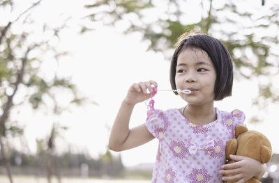 Girl blowing bubbles while holding teddy bear in park