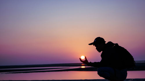 Optical illusion of silhouette man holding sun while standing at beach against orange sky during sunset