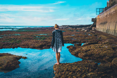 Rear view of woman standing on rock by sea against sky