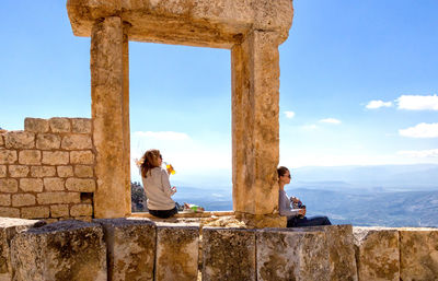 Women having food and drink while sitting on old ruin against sky