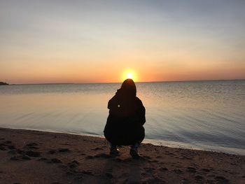 Rear view of silhouette woman crouching on shore at beach during sunset