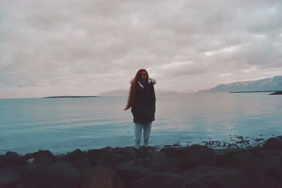 Woman standing on rocky shore against cloudy sky