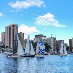 Sailboats in river by buildings against cloudy sky