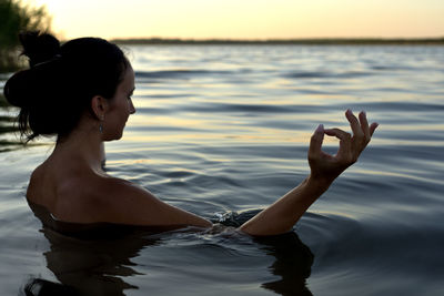 Shirtless woman meditating in sea during sunset