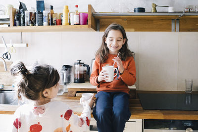 Smiling girl having drink while sister holding mug in kitchen at home