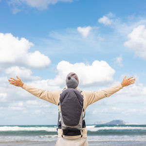 Rear view of man with arms raised standing at beach against sky