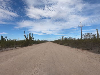 Dirt road amidst electricity pylon against sky