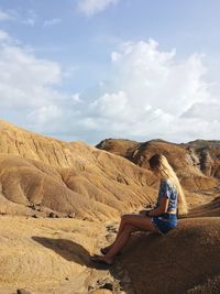 Side view of young woman sitting on rock against sky at desert