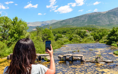 Rear view of girl holding phone taking photos of beautiful river and nature in summer