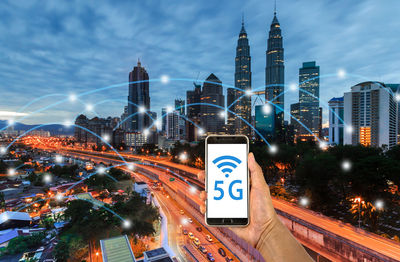 Digital composite image of man holding mobile phone with computer icons against cityscape at dusk