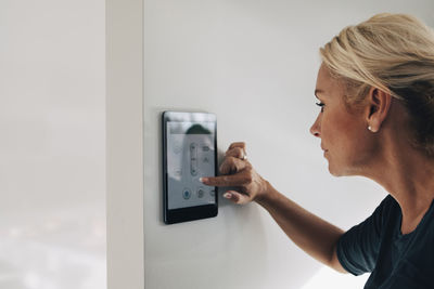 Blond woman adjusting thermostat using digital tablet mounted on white wall at home