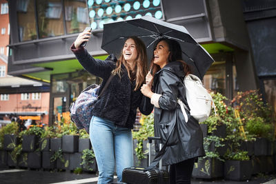 Happy friends with umbrella taking selfie through mobile phone while standing in city