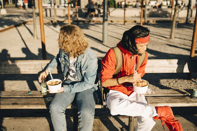 Friends eating take out food while sitting on bench in city during sunny day