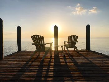 Empty chairs and table on pier over sea against sky during sunset