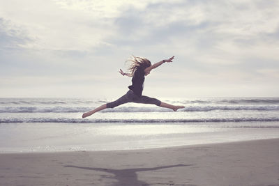 Carefree girl jumping at beach against cloudy sky