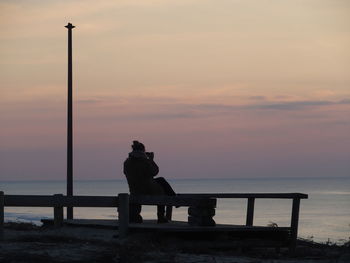 Man sitting on bench by sea against sky during sunset
