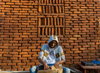 Full length of man sitting against brick wall playing a drum