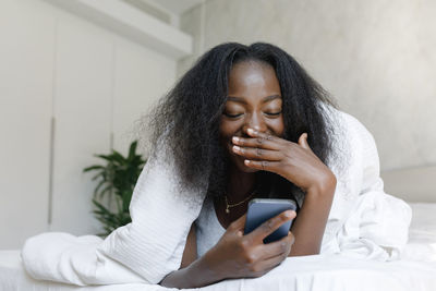 Happy young woman looking at mobile phone lying on bed