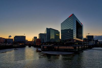 Modern buildings by river against sky during sunset