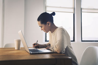 Businesswoman writing while sitting with laptop computer at desk in office