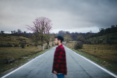 Side view of anonymous blurred man in casual wear walking on empty asphalt road among green fields with cloudy sky on background