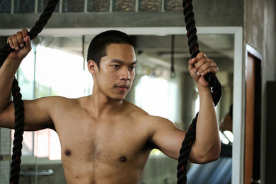 Shirtless young man holding ropes while standing in gym