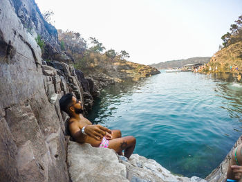 Full length of shirtless man relaxing on rock against sky