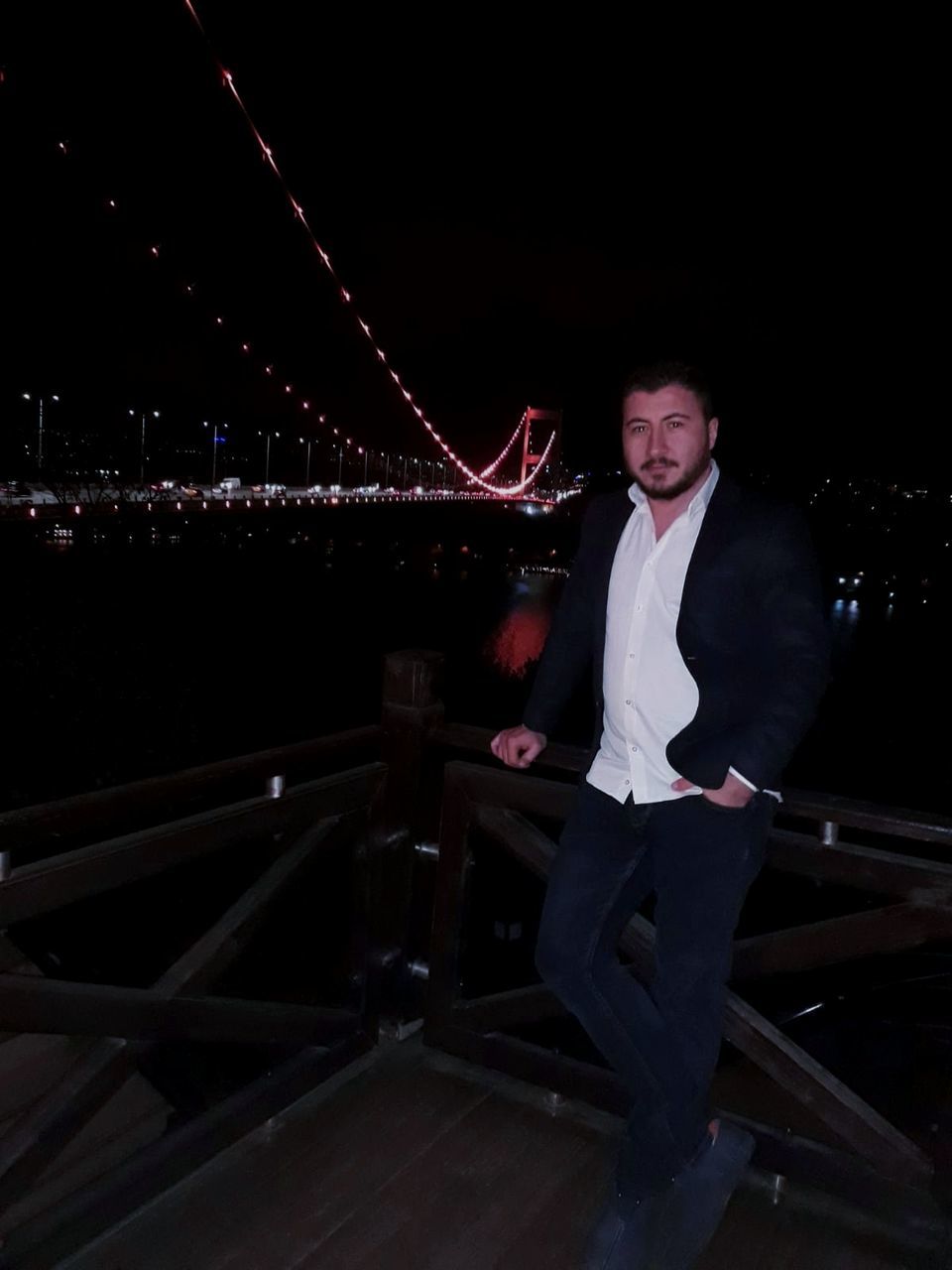 night, one person, real people, illuminated, full length, architecture, young adult, front view, built structure, leisure activity, casual clothing, young men, lifestyles, sitting, looking, connection, transportation, bridge, railing, bridge - man made structure, outdoors