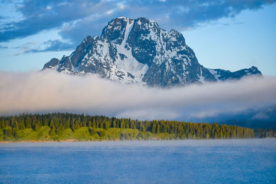 Scenic view of snowcapped mountains, fog, lake, and trees against sky