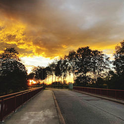 Empty road by silhouette trees against sky during sunset