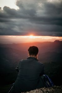 Rear view of man sitting on cliff against cloudy sky during sunset