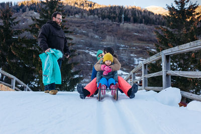Standing father talking with three daughters sitting on one sled