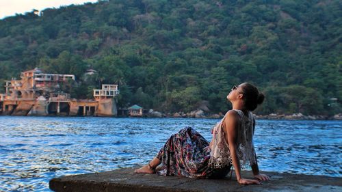 Woman sitting on retaining wall by lake against trees