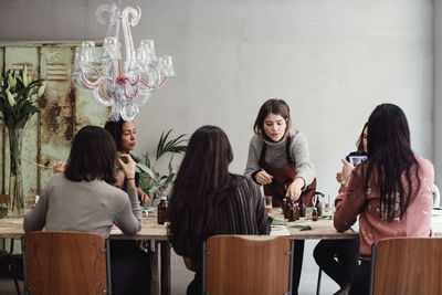 Confident female entrepreneur preparing perfume while standing amidst colleagues sitting at table in workshop