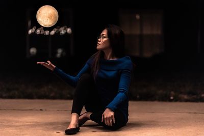 Young woman looking away while sitting on illuminated lamp