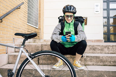 Full length happy delivery woman in uniform sitting on steps near bike and browsing cellphone while taking break during work