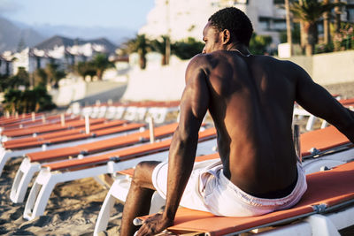 Rear view of shirtless man sitting on deck chair at beach