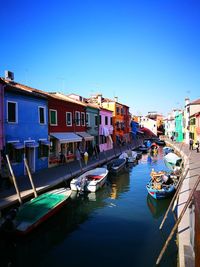 Boats moored in canal by buildings against clear blue sky