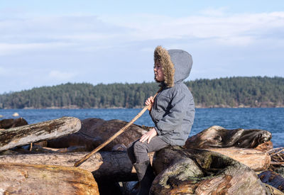 Side view of boy wearing warm clothing sitting on logs by sea against sky