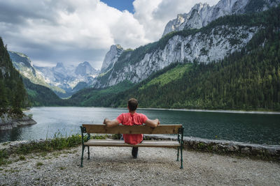 Rear view of man sitting on bench by lake against mountains