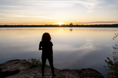 Rear view of woman standing at lakeshore against sky during sunset