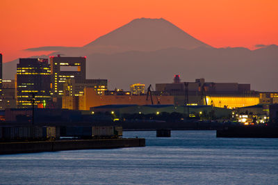 Illuminated city by sea against sky at sunset