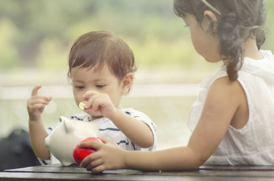 Baby boy with sister inserting coin in piggy bank