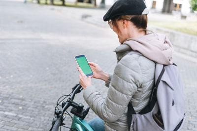 Portrait of young woman in cap and sunglasses using mobile on bicycle on sunny autumn day in city 
