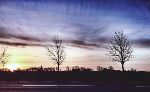 Bare trees on landscape against cloudy sky at sunset