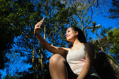Low angle view of woman taking selfie while sitting in forest