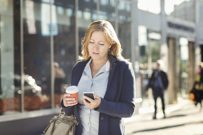 Businesswoman using mobile phone and holding disposable coffee cup while walking on sidewalk