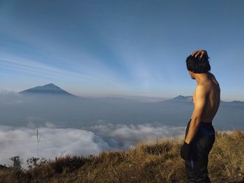 Side view of shirtless man standing on cliff against blue sky