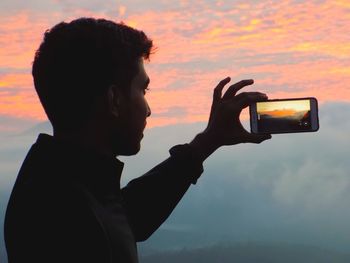 Side view portrait of man photographing camera against sky during sunset