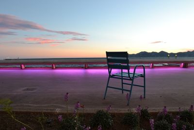 Empty chair on illuminated purple promenade against sky during sunset